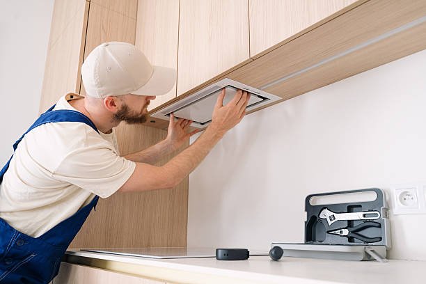 A professional handyman is focused on installing a range hood above the kitchen cabinetry in an apartment. Tools are neatly organized on the countertop, showcasing a tidy workspace.