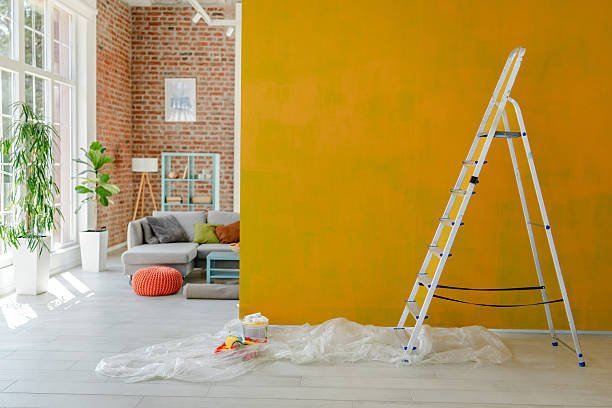 Ladder placed beside a vibrant yellow wall mid-paint, with a comfy living space and decorative shelving visible behind.